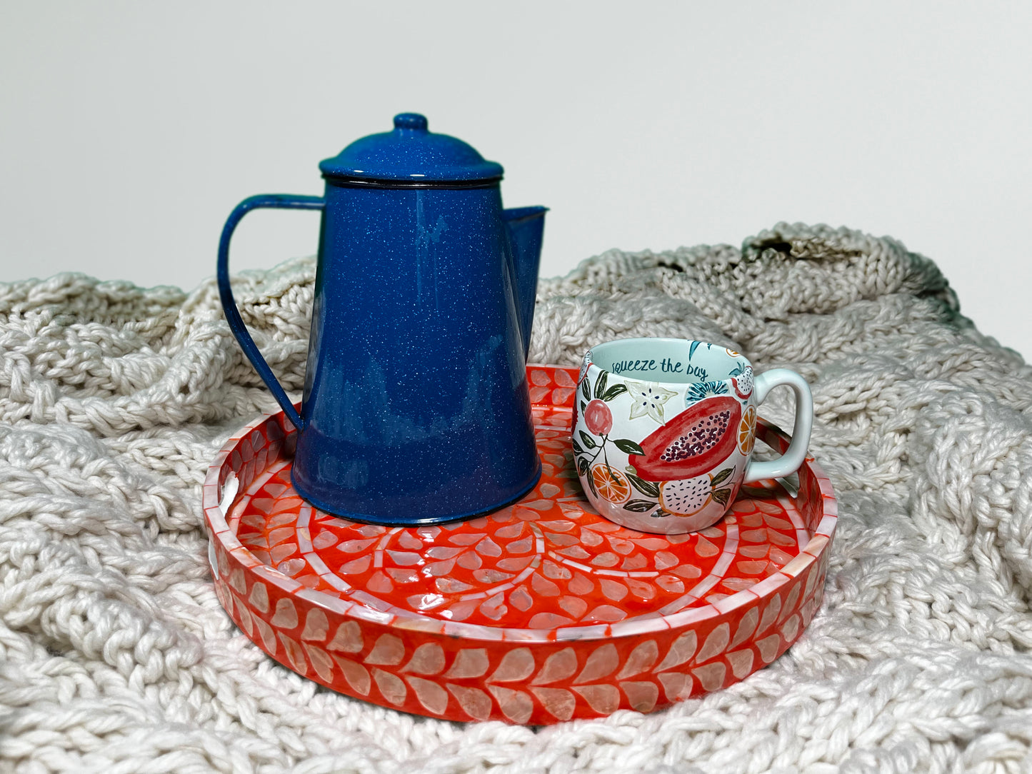 A Deep Coral Mother of Pearl Tray on a beige blanket that has blue water kettle and a fruit pattern cup 