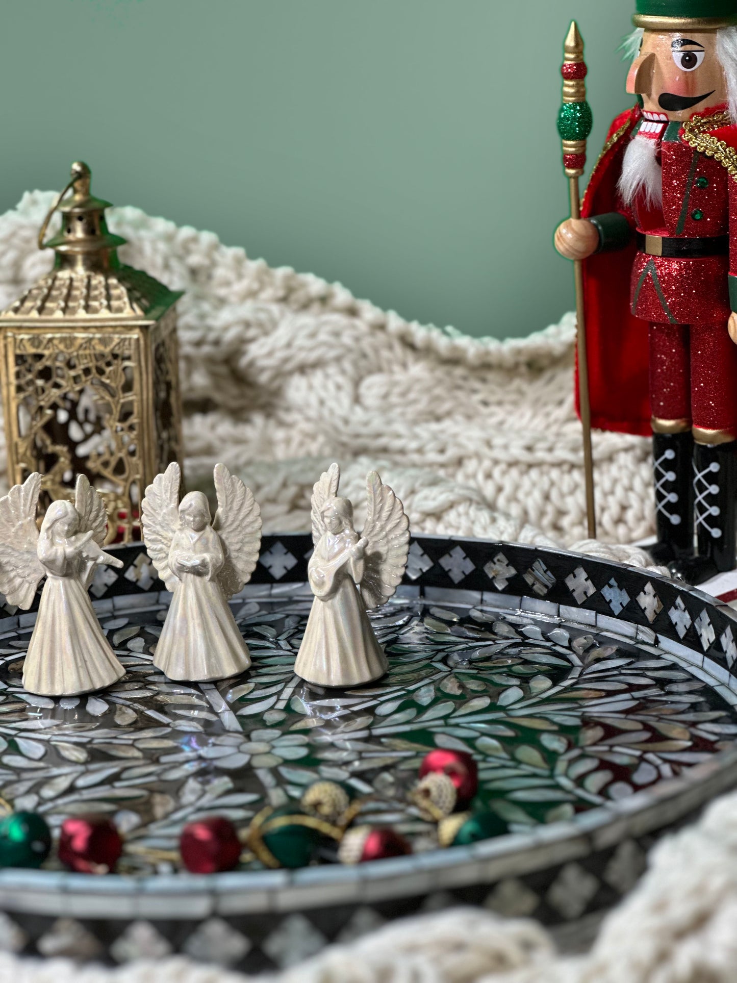 close up shot of the Black Sea Mother of Pearl Tray on a blanket, with three angels and Christmas bells on, and a gold candle holder, a Christmas nut cracker in the back