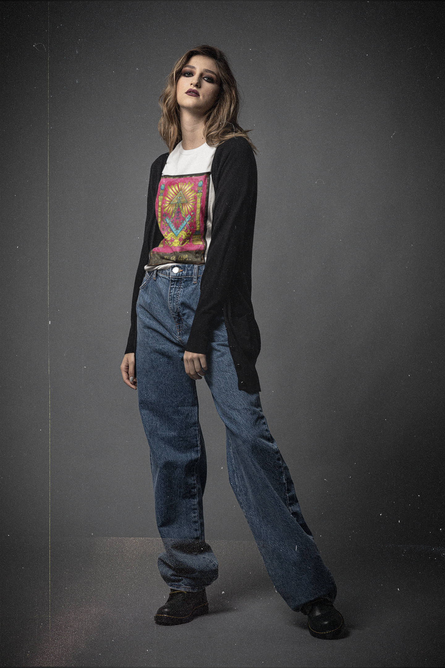 White blond woman in a white ace of cups tarot card t-shirt, black cardigan, blue jeans, and black shoes posing in a black background studio