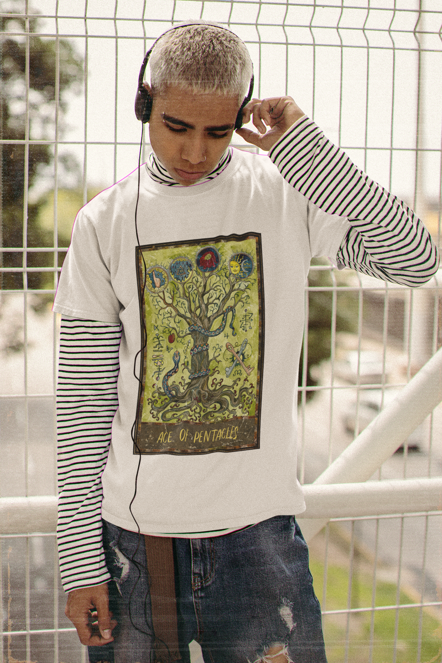 A young man standing in front of white fences, listening to music wearing a long sleeved black and white stripe shirt, a white Ace of Pentacles Tarot Card T-shirt, ripped blue jeans, and brown belt