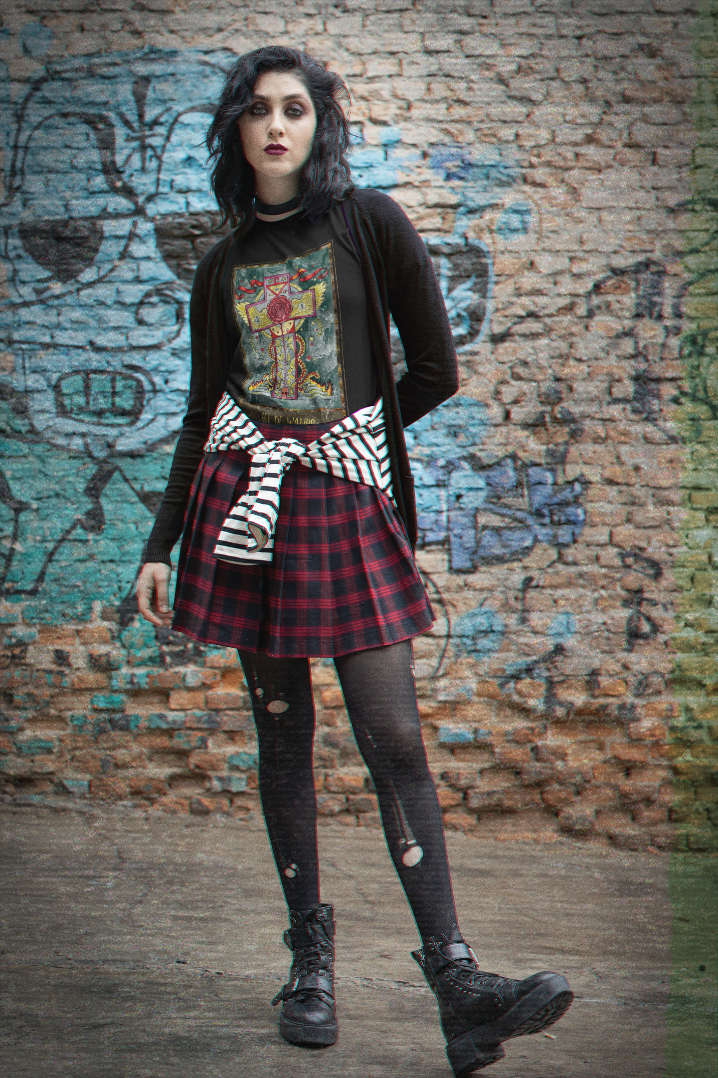 A woman wearing a black Ace of Wands Tarot Card T-shirt, black cardigan, checkered black and red skirt, black and white striped shirt tying around her waist ripped black stockings, and black boots