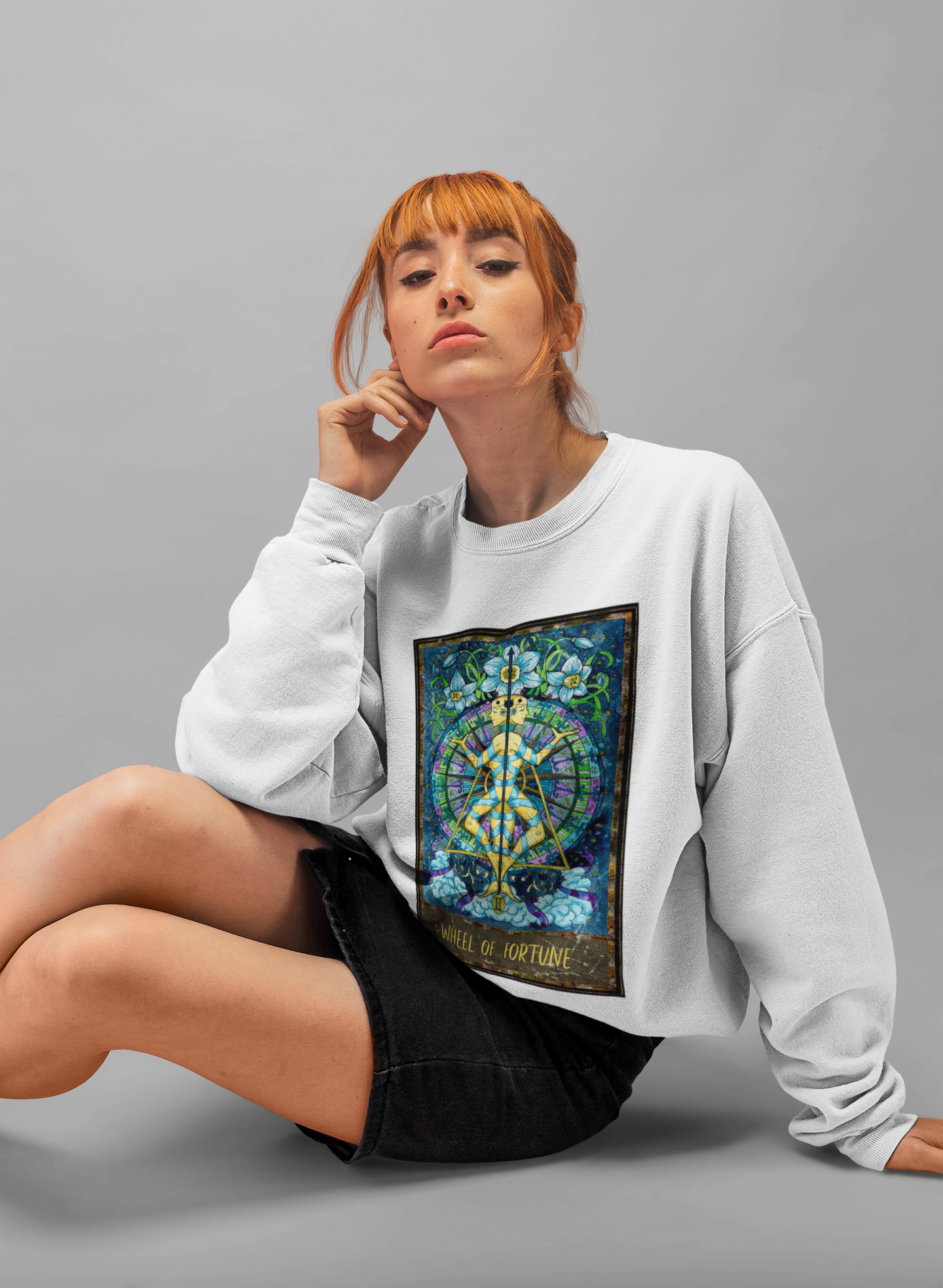 A woman sitting in a studio wearing a white Wheel of Fortune Tarot Card Sweatshirt and a black skirt