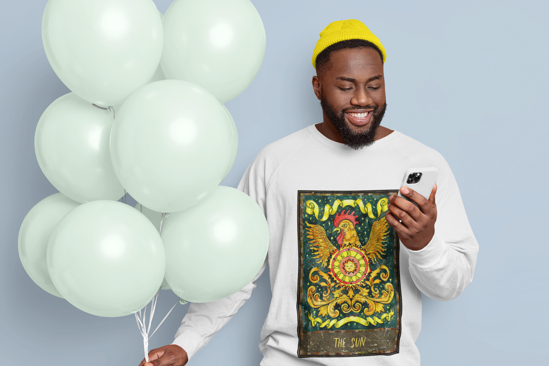 A smiling man holding balloons and looking at his phone wearing a white The Sun Tarot Card Sweatshirt and a yellow beanie