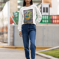 A woman with long hair posing on a street wearing a white Justice - Tarot Card Sweatshirt, blue jeans and brown boots