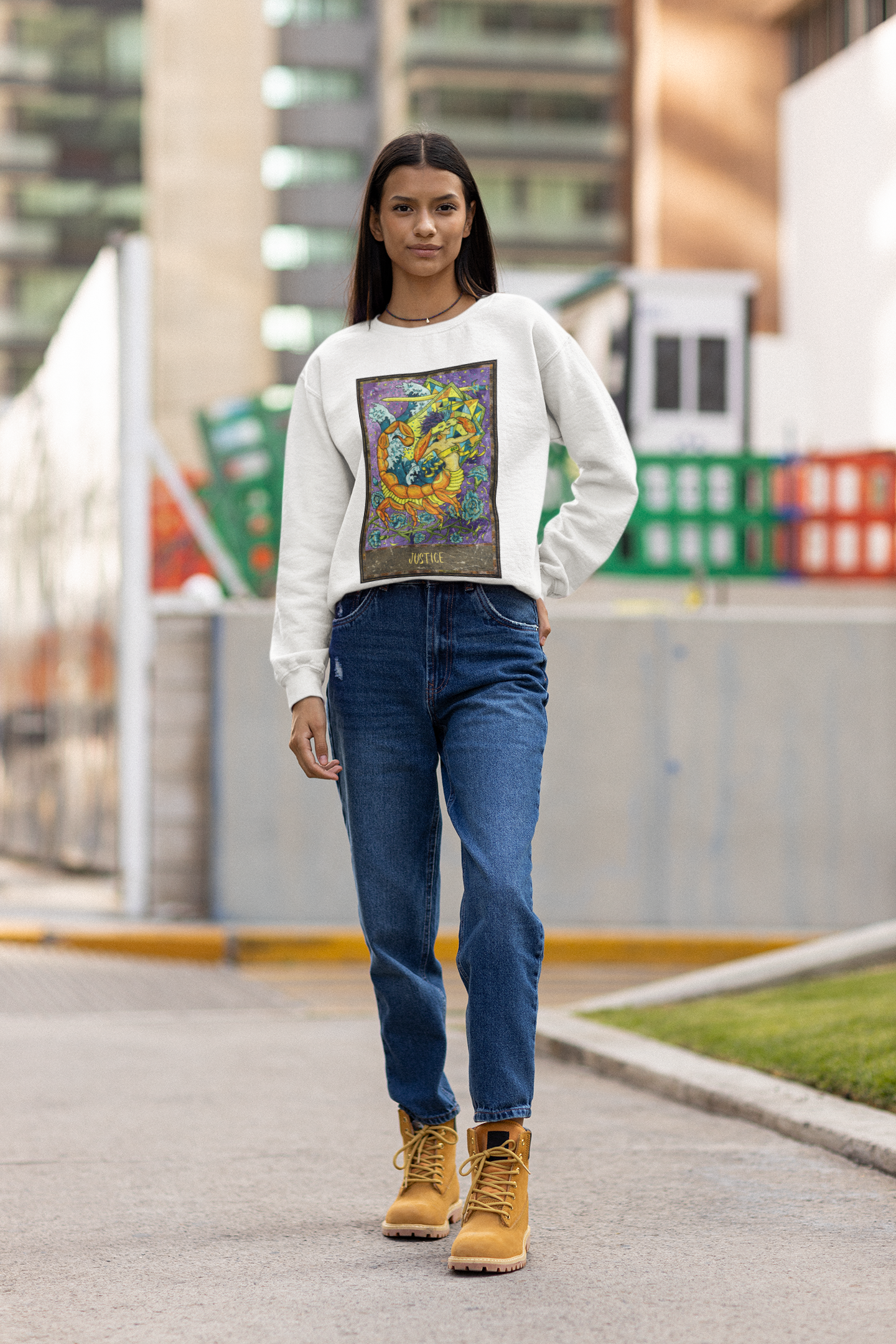 A woman with long hair posing on a street wearing a white Justice - Tarot Card Sweatshirt, blue jeans and brown boots