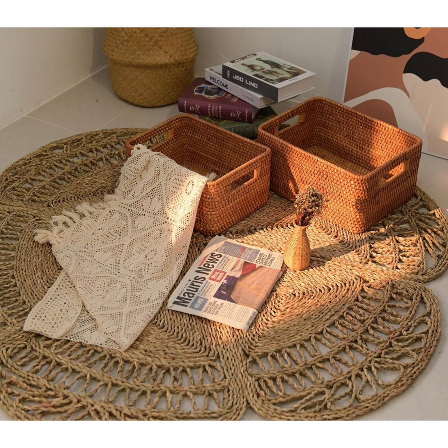 Flower Seagrass Rug in the corner of a room with baskets, a newspaper, a cloth and a small flower vase on top, there are some books and a painting in the background