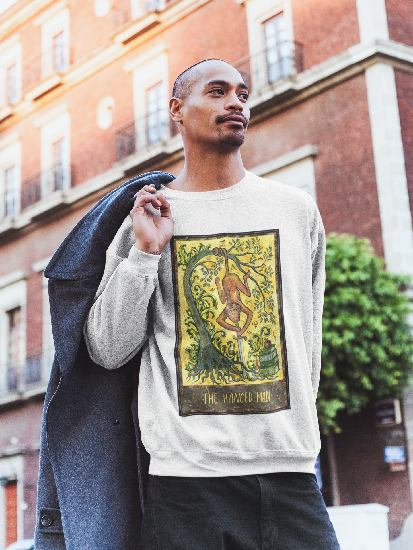 A man wearing a white The Hanged Man Tarot Card Sweatshirt, black pants holding a coat