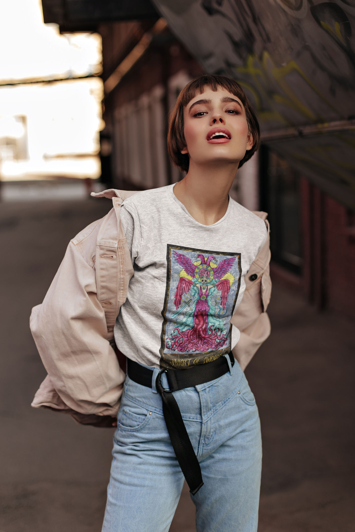 A short haired woman posing wearing a white Knight of Swords Tarot Card T-shirt, pink jacket, and blue jeans