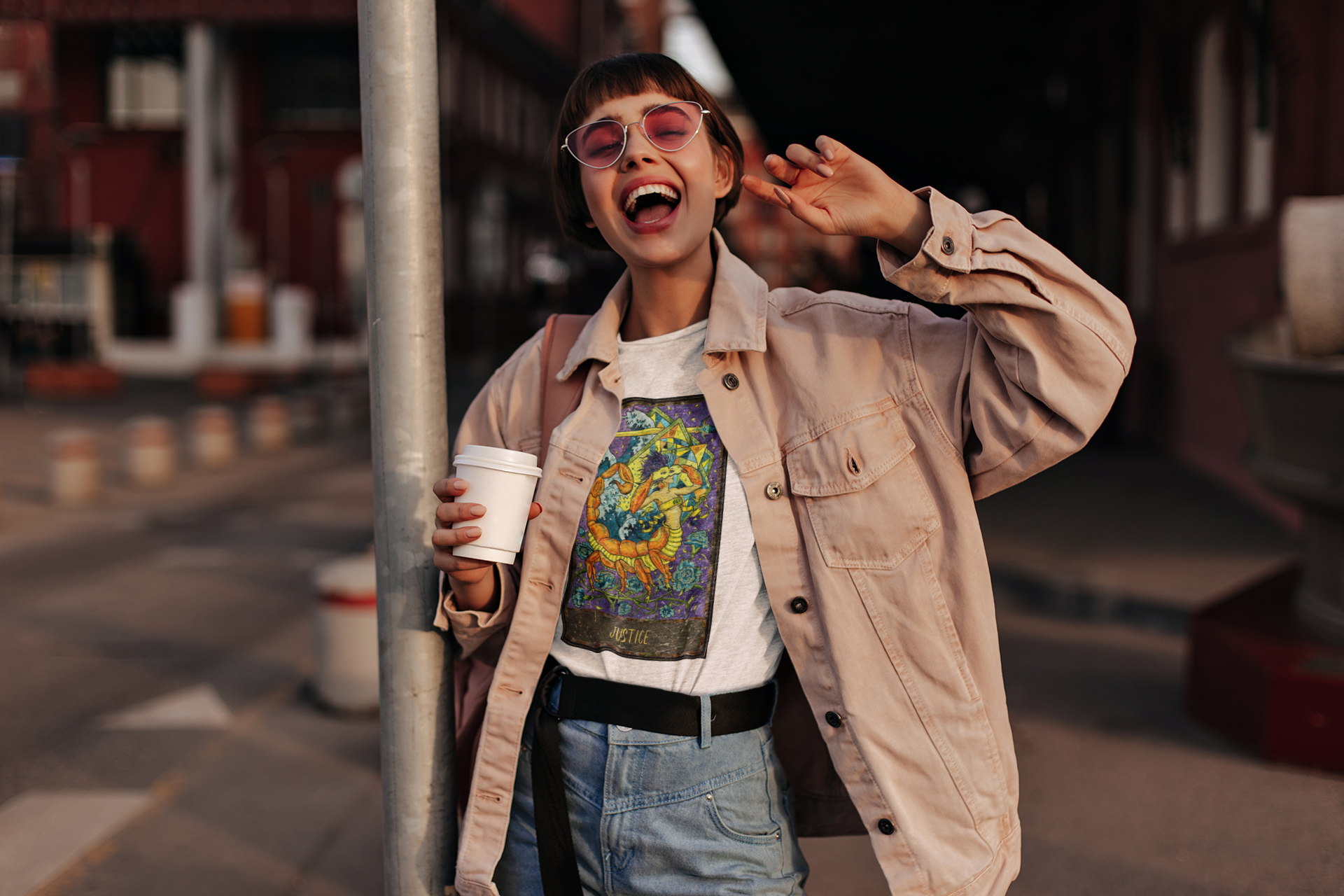 A woman with sunglasses holding a coffee cup wearing a white Justice - Tarot Card T-shirt, pink denim jacket, and blue jeans