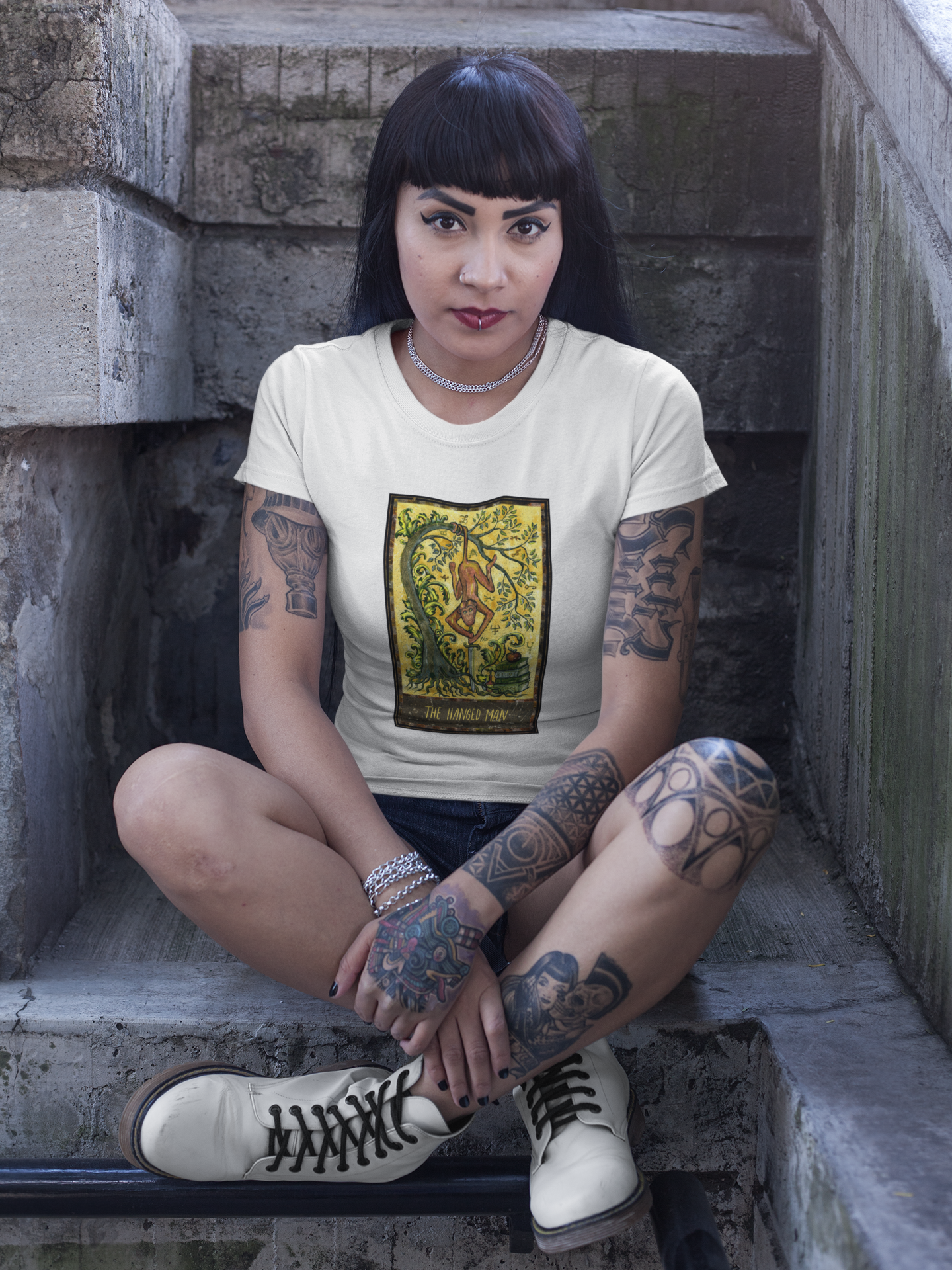 A woman with tattoos wearing a white The Hanged Man Tarot Card T-shirt sitting outside a church