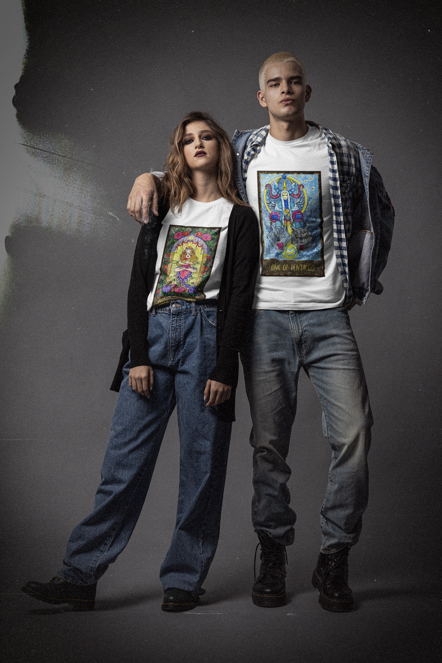 A man and woman posing in a studio: the man is wearing a white King of Pentacles Tarot Card T-Shirt inside a collar shirt and denim jacket, fade blue jeans, black boots; the woman is wearing a white Queen of Pentacles Tarot Card T-Shirt inside a black cardigan, blue jeans, and black shoes