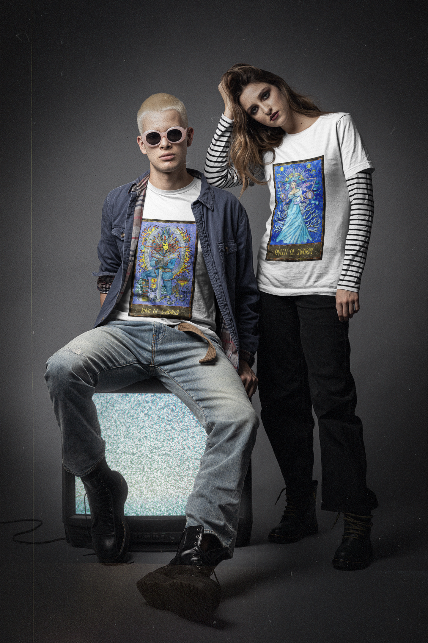 A man sitting down wearing a white King of Swords Tarot Card T-Shirt inside a dark blue shirt, washed blue jeans, and black boots. A woman standing next to him wearing a white Queen of Swords Tarot Card T-Shirt outside of a stripes inner shirt, black pants, and black boots