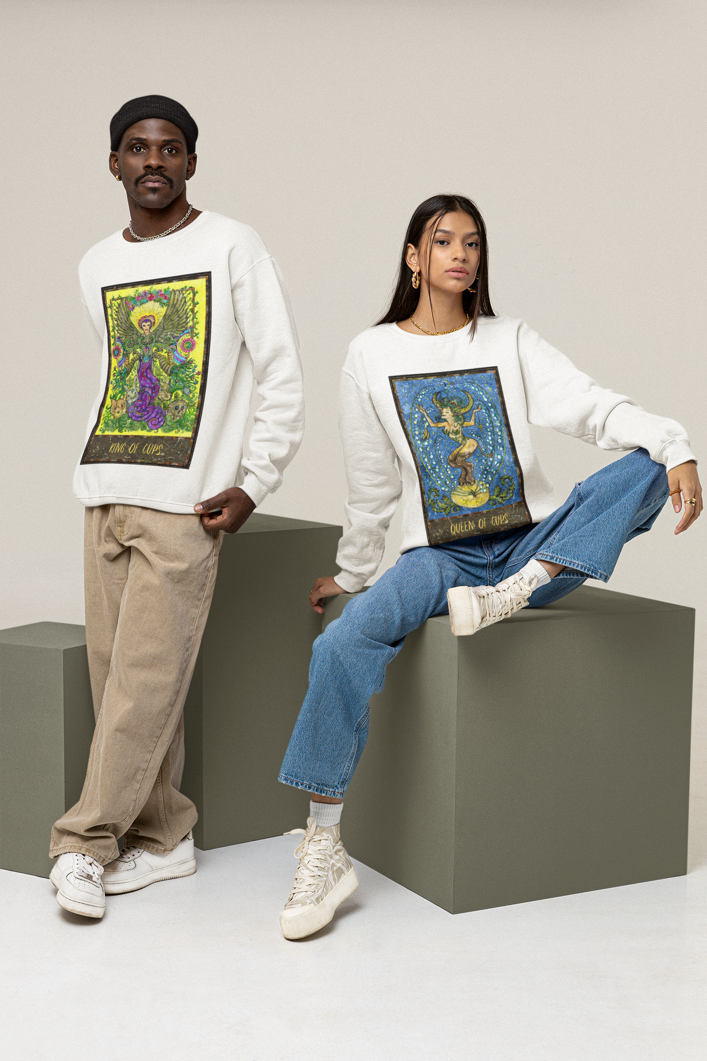 A woman wearing a white Queen of Cups Tarot Card Sweatshirt with blue jeans, and white shoes. Standing next to her is a man wearing a white King of Cups Tarot Card Sweatshirt with beige pants and white shoes