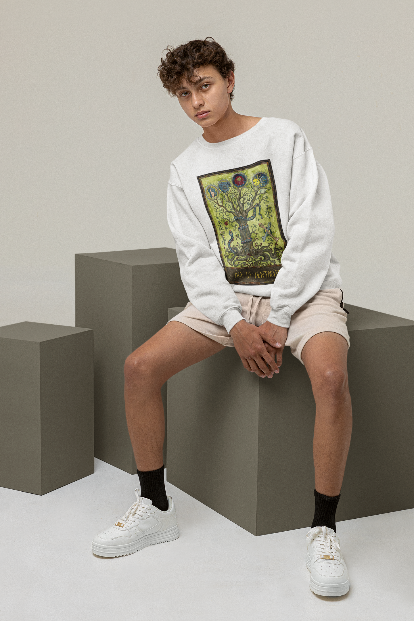 A young man with curly hair wearing a white Ace of Pentacles Tarot Card Sweatshirt, beige shorts, black socks, and white shoes sitting on a a grey block