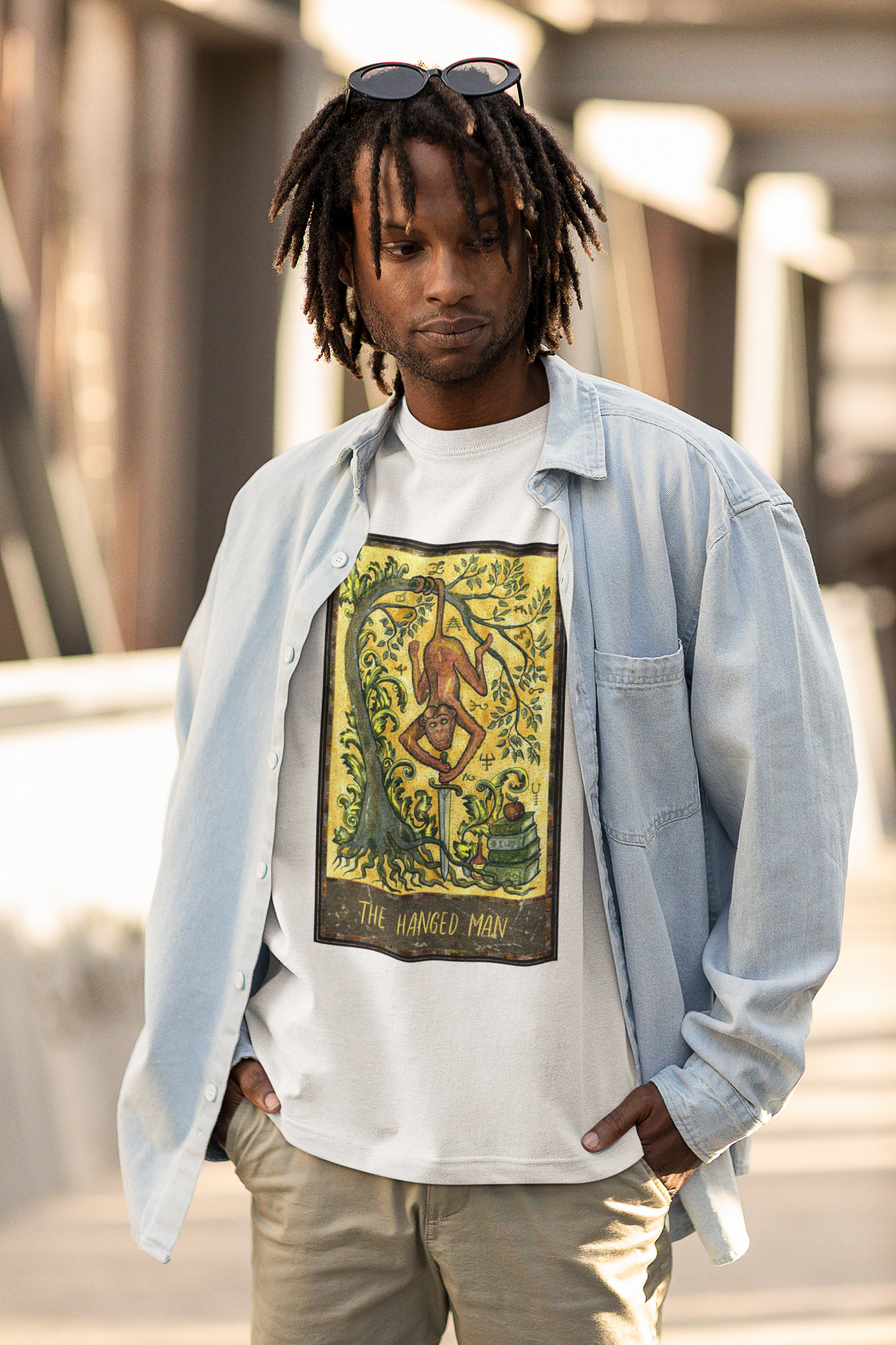 A man with locs and sunglasses wearing a white The Hanged Man Tarot Card T-shirt inside a light blue shirt, beige pants