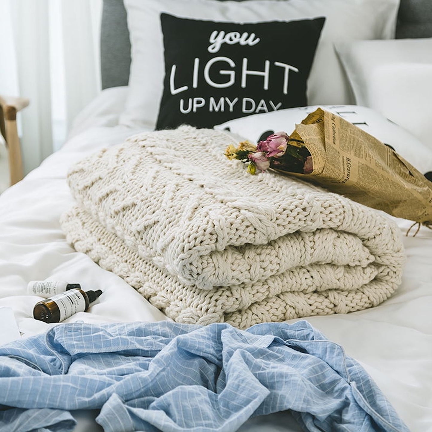 Beige Knitted Throw Blanket folded on a bed with pillows, a bouquet of flowers, two unknown skin care bottle, and a blue cloth