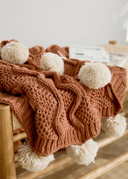 close up shot of the rusty orange Pompom Fringes Solid Chenille Knitted Throw Blanket on a shelf