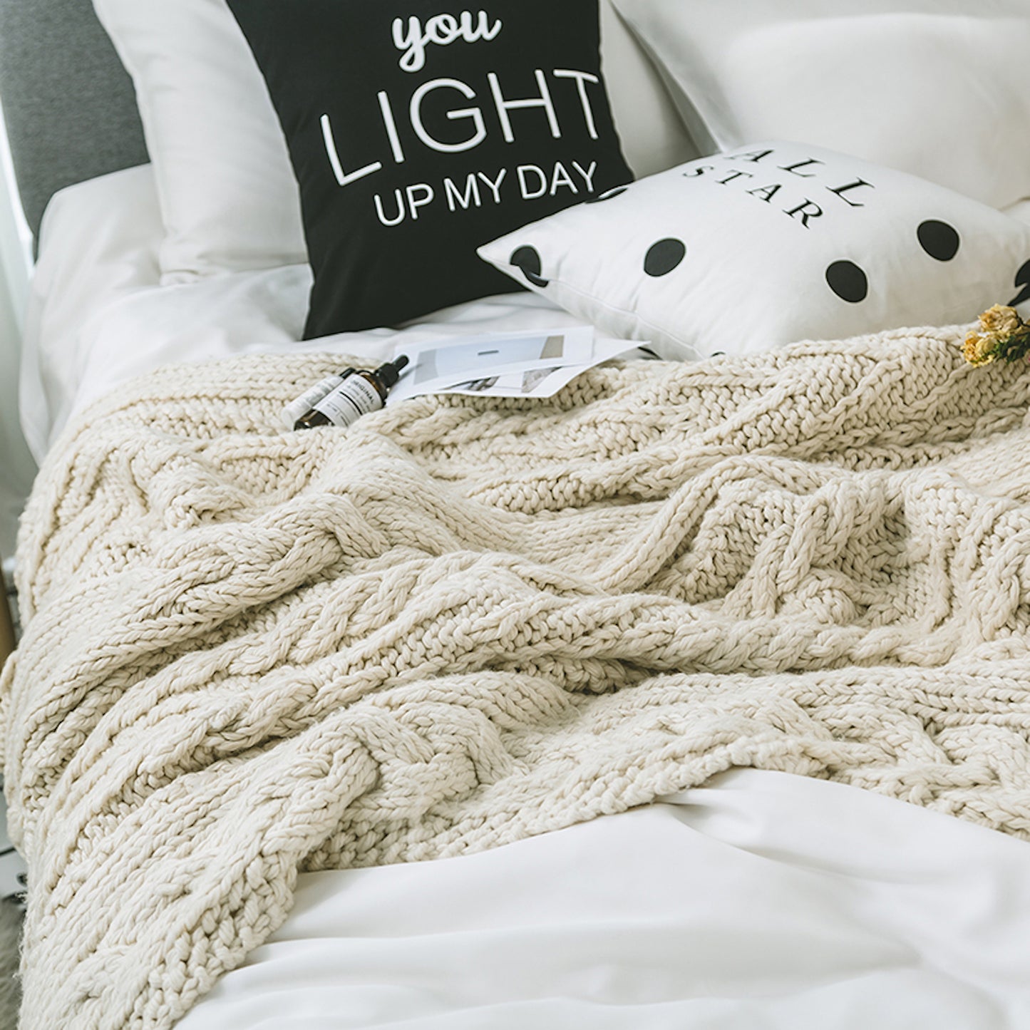 Beige Knitted Throw Blanket spreading out on a bed with pillows and skin care bottles