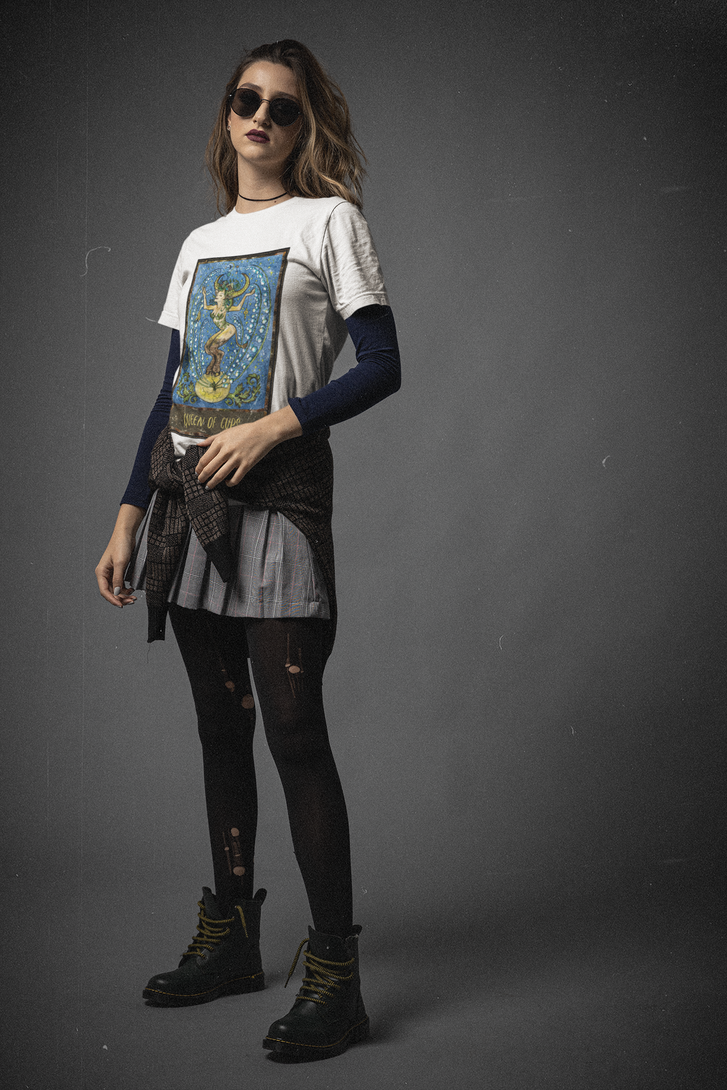 A woman wearing black sunglasses, a white Queen of Cups Tarot Card T-Shirt, black inner shirt, a grey skirt with ripped black stockings and black boots 