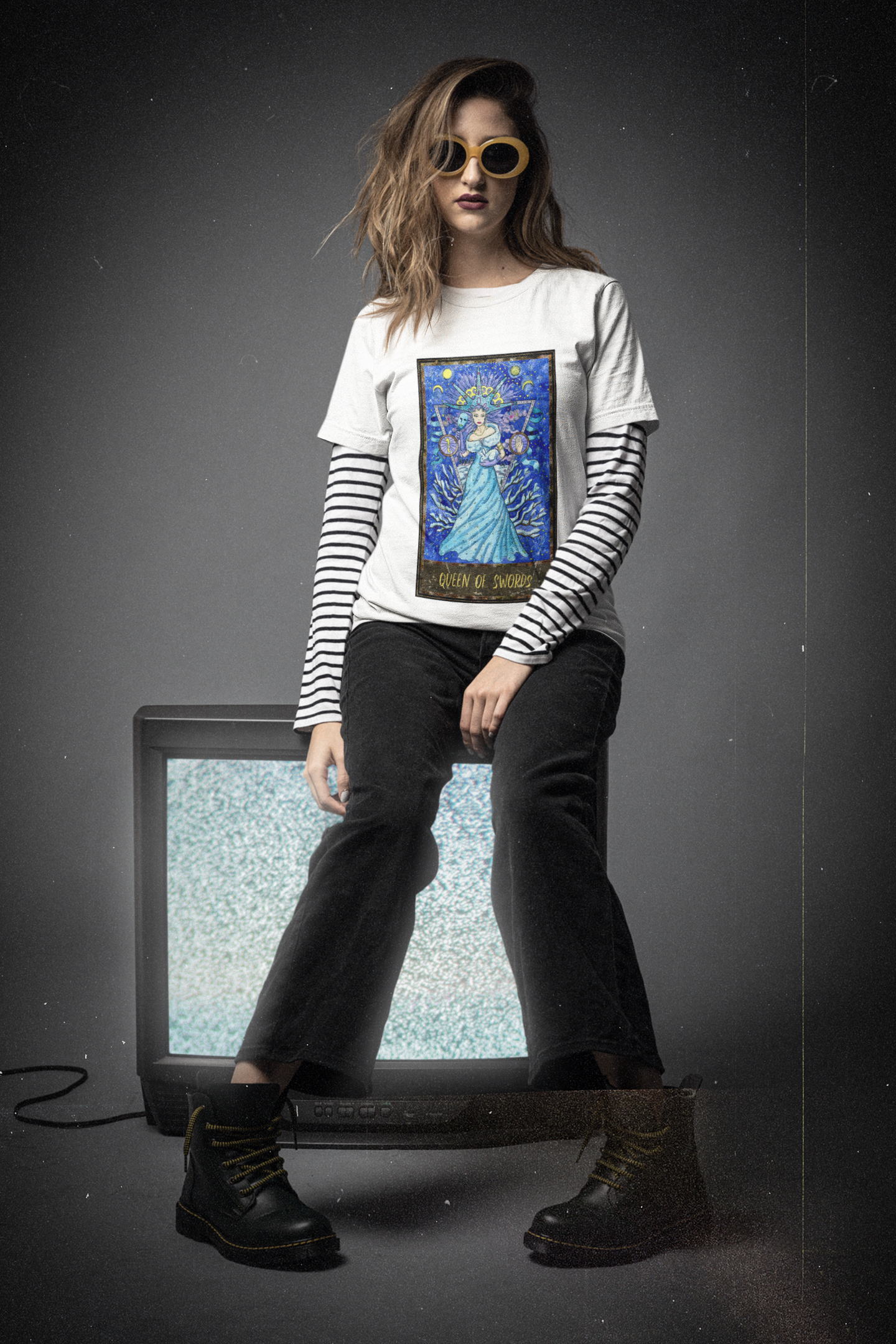 A woman wearing a white Queen of Swords Tarot Card T-Shirt over a striped long sleeves, black pants and black boots