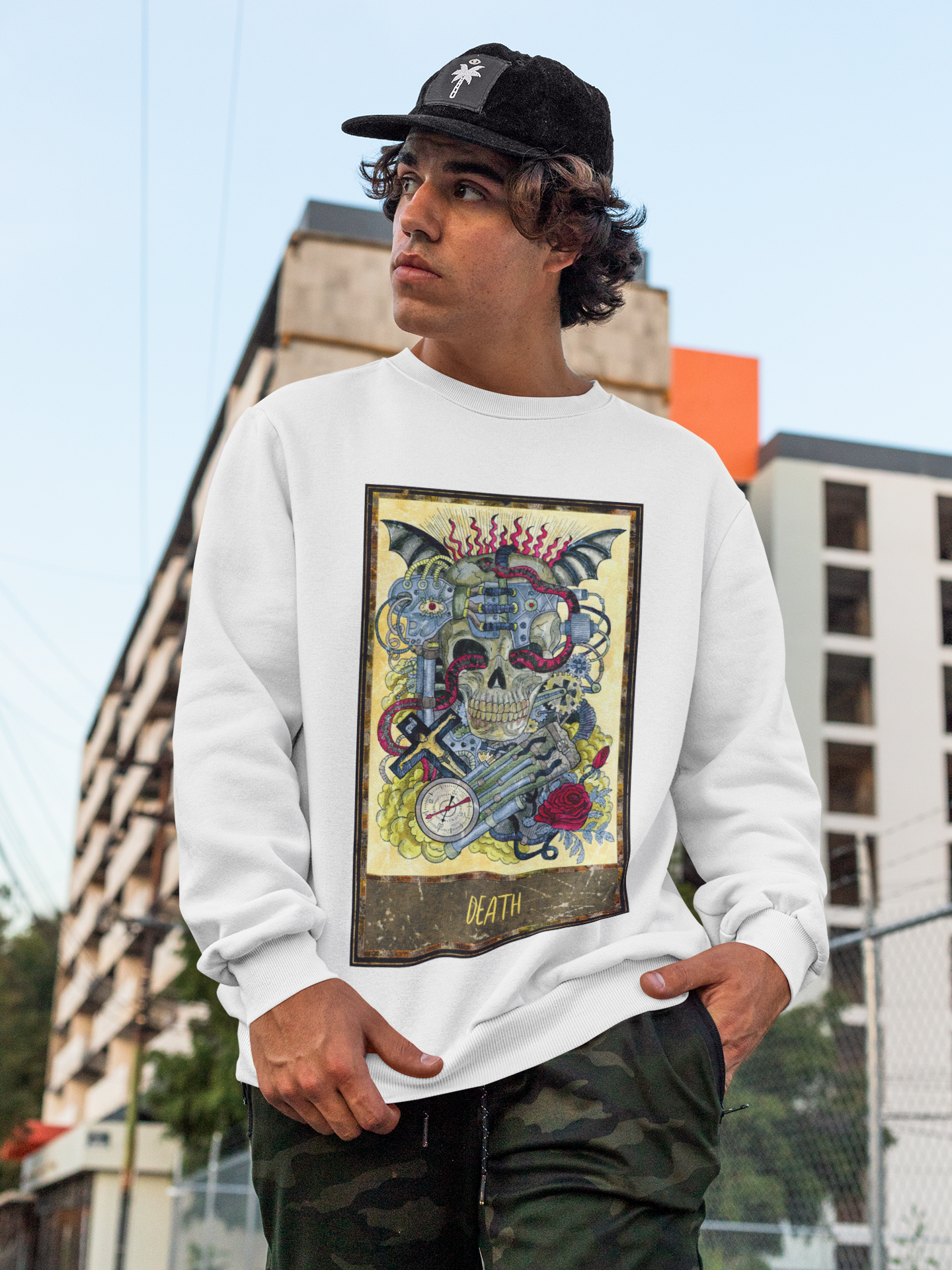 A curly haired man with a cap wearing a white Death Tarot Card Sweatshirt and cargo pants