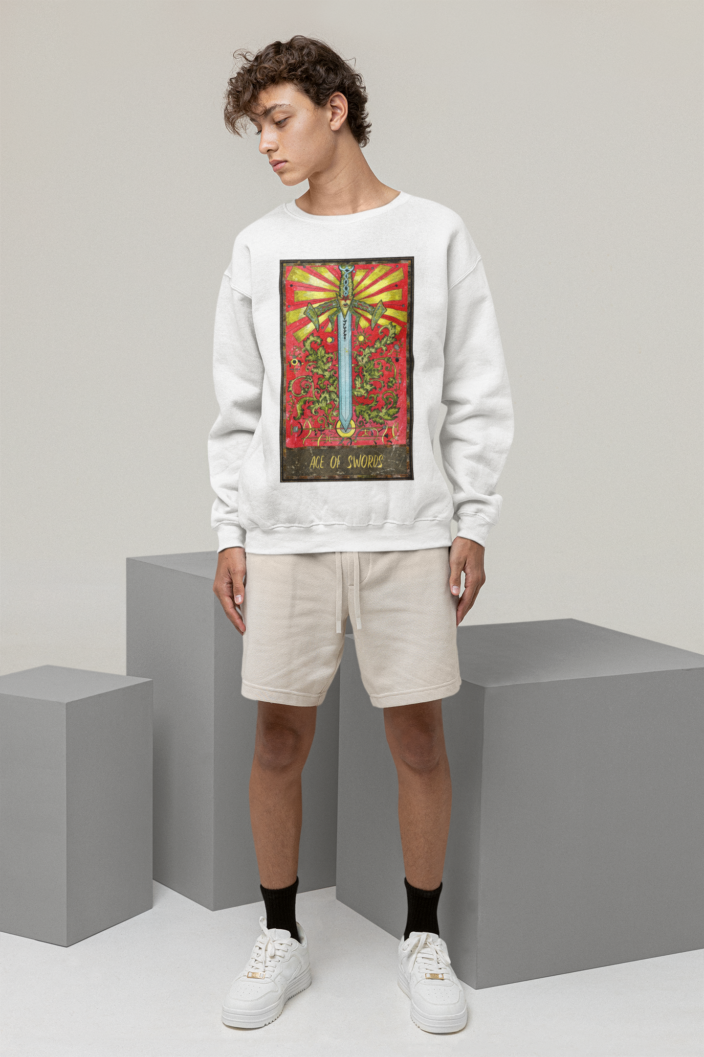 A young man standing in a studio wearing a white Ace of Swords Tarot Card sweatshirt, beige shorts, black socks, and white shoes