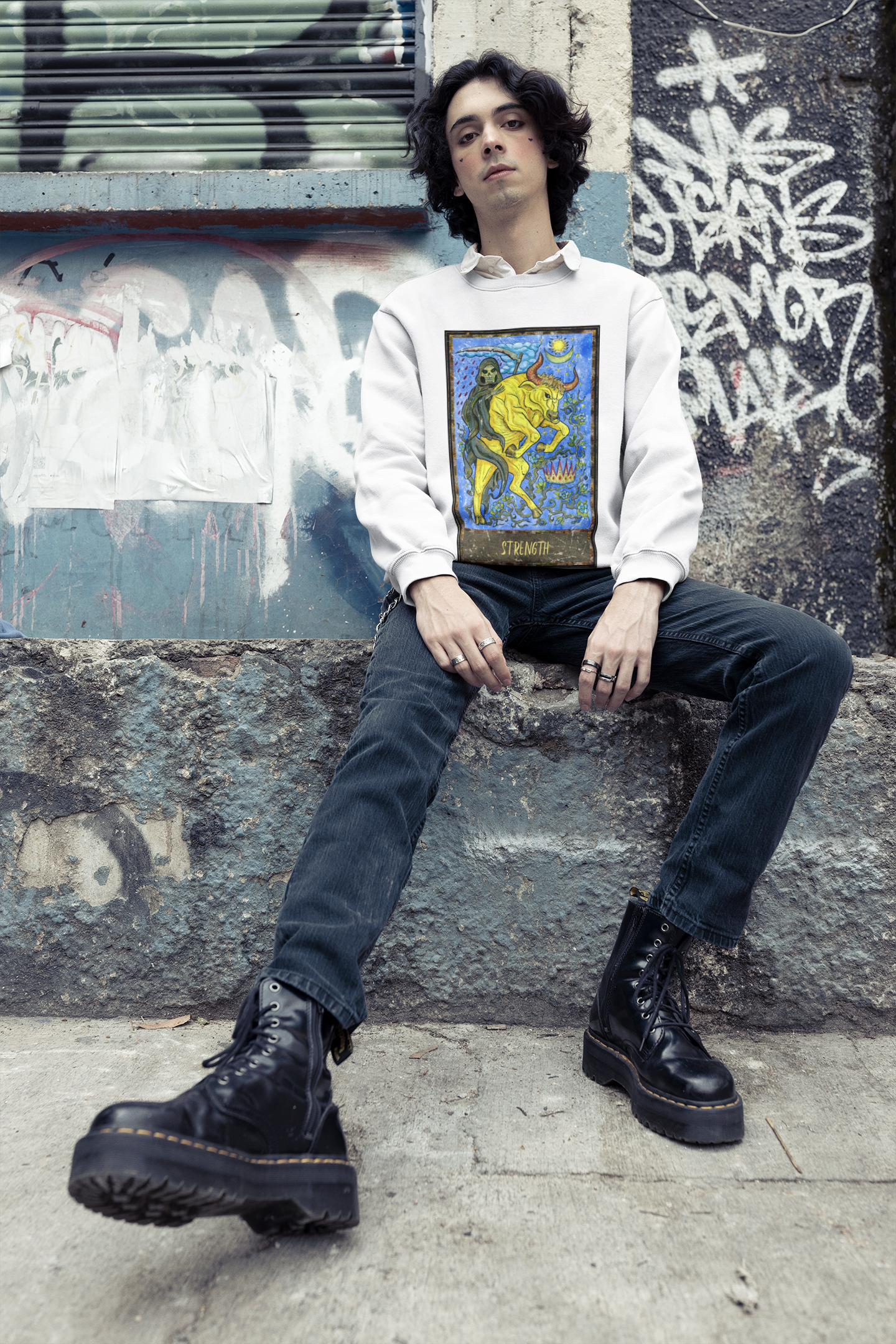 A young boy wearing a white Strength Tarot Card Sweatshirt over a white collar shirt, black jeans and black boots