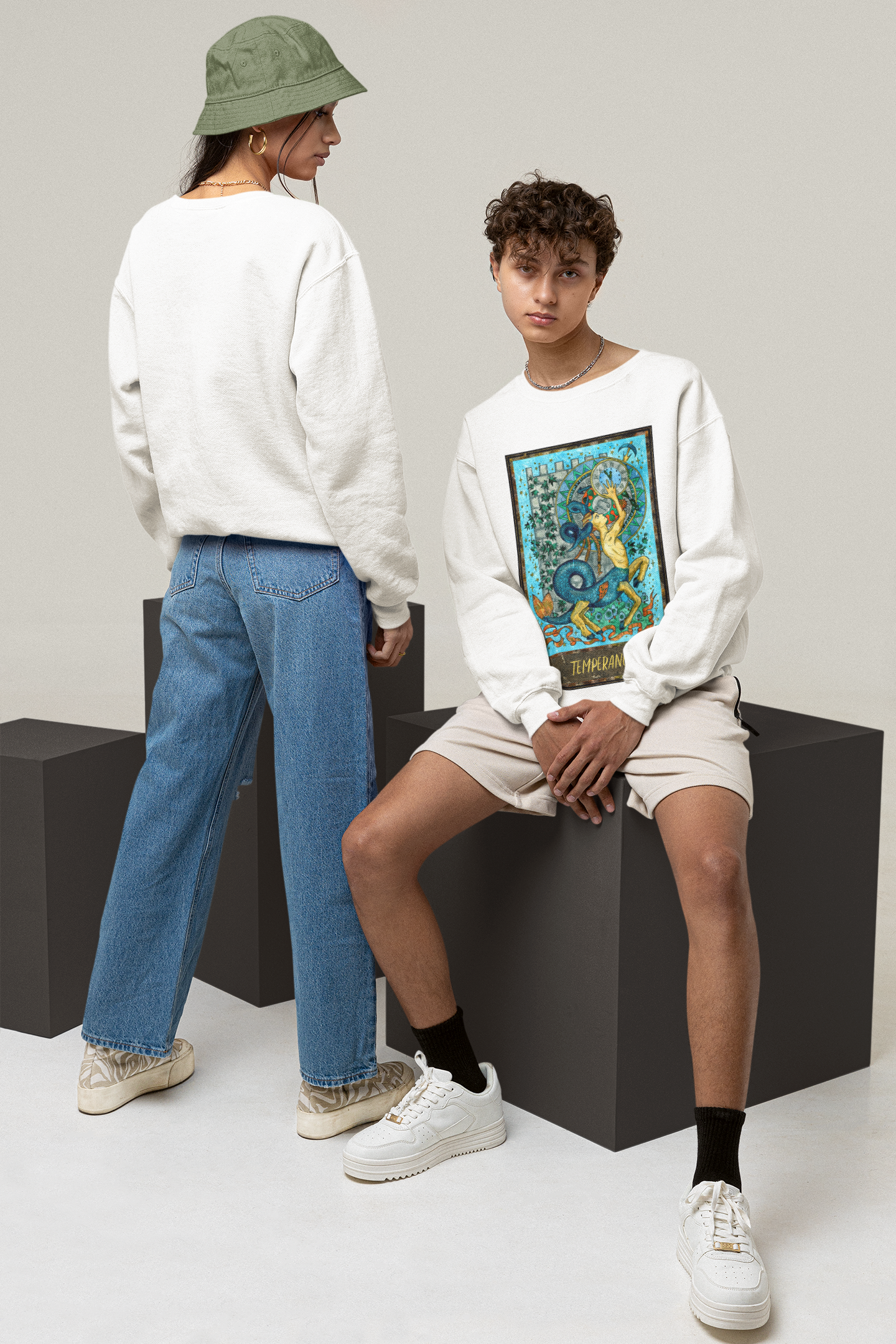 A young man wearing a white Temperance Tarot Card Sweatshirt, beige shorts, and white shoes sitting on a block. A woman wearing a white sweatshirt, blue jeans and a green bucket hat