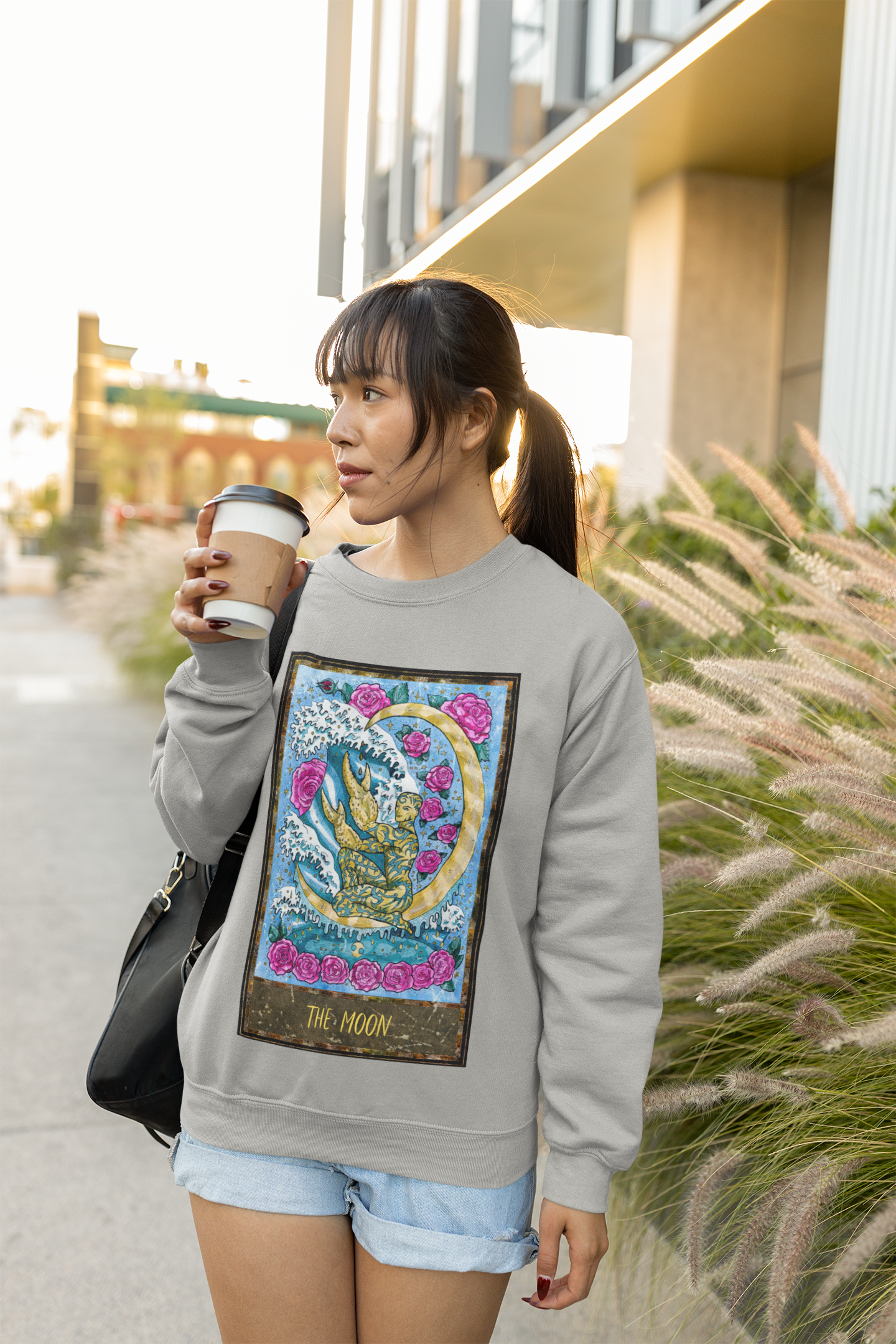 A woman drinking coffee on the street wearing a grey The Moon Tarot Card Sweatshirt and denim shorts