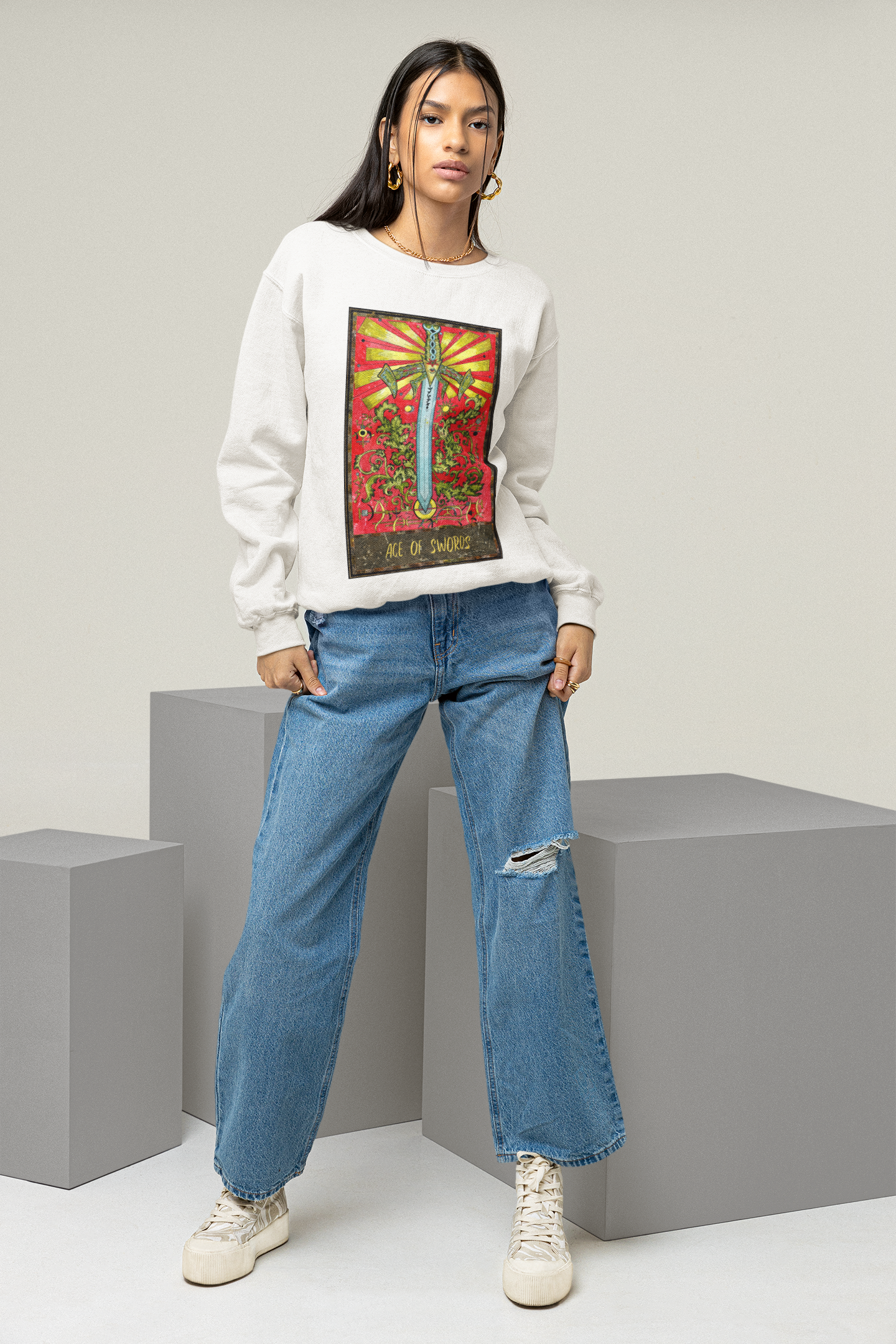 A young woman posing in a studio, wearing a white Ace of Swords Tarot Card Sweatshirt, ripped blue jeans, and white shoes