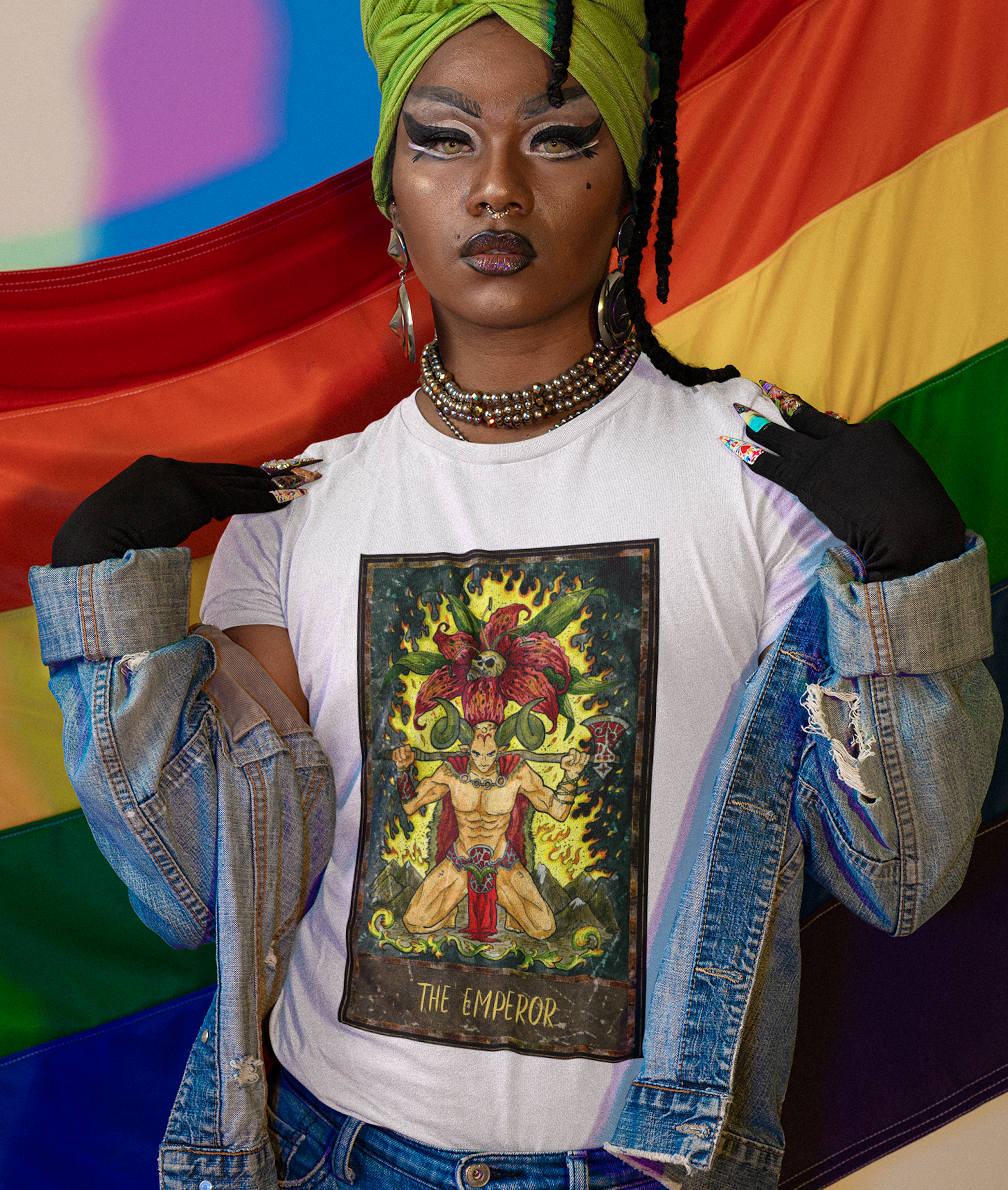 A person posing in front of the pride flag wearing a white The Emperor Tarot Card T-shirt and blue denim jacket, and blue jeans