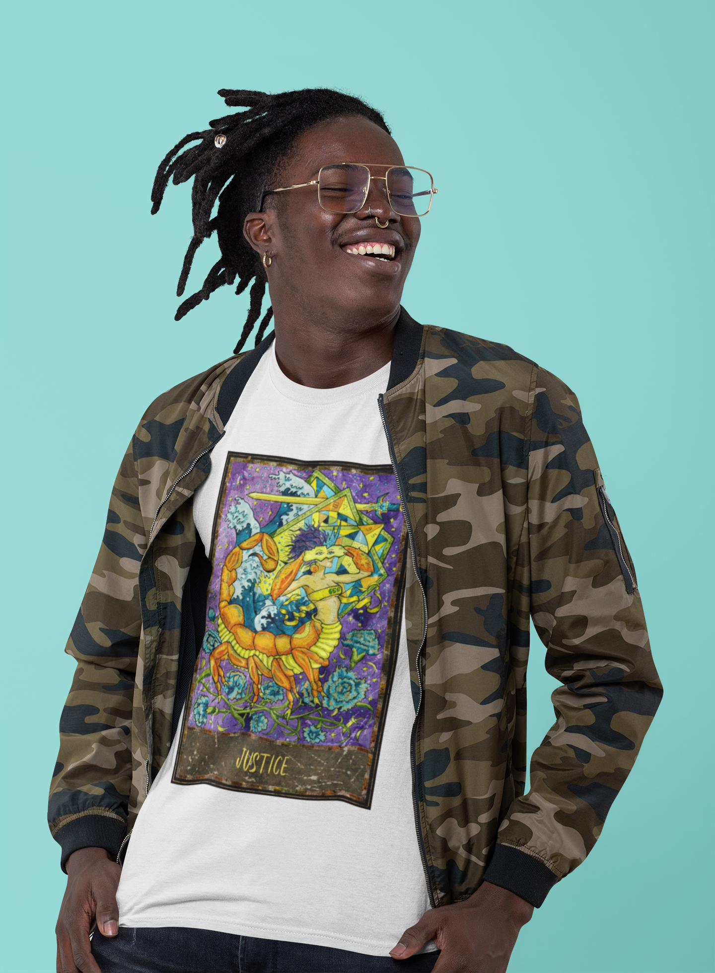 A smiling man with retro glasses wearing a white Justice - Tarot Card T-shirt inside a camo jacket and jeans