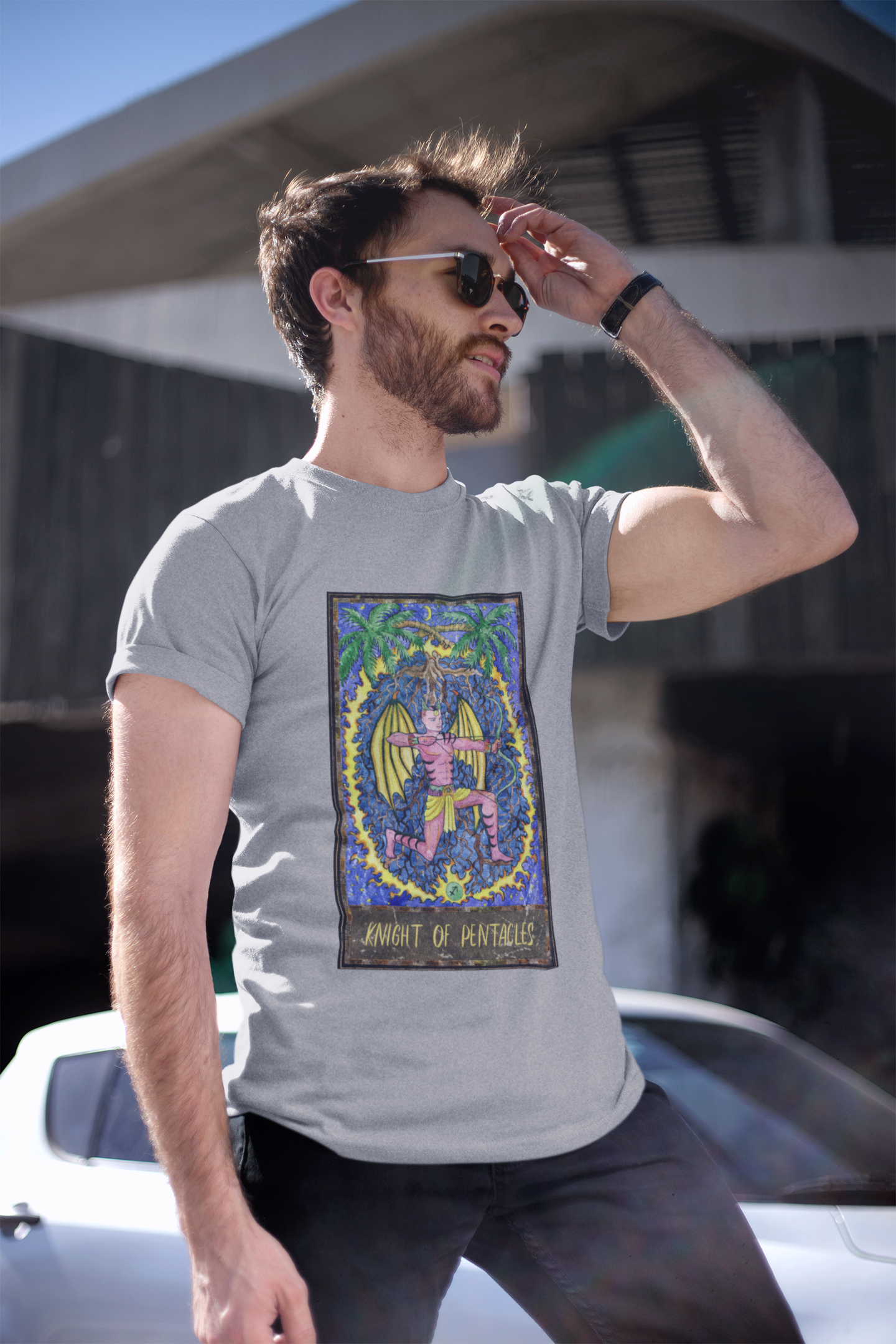 A man wearing sunglasses, a grey Knight of Pentacles Tarot Card T-shirt, and black pants