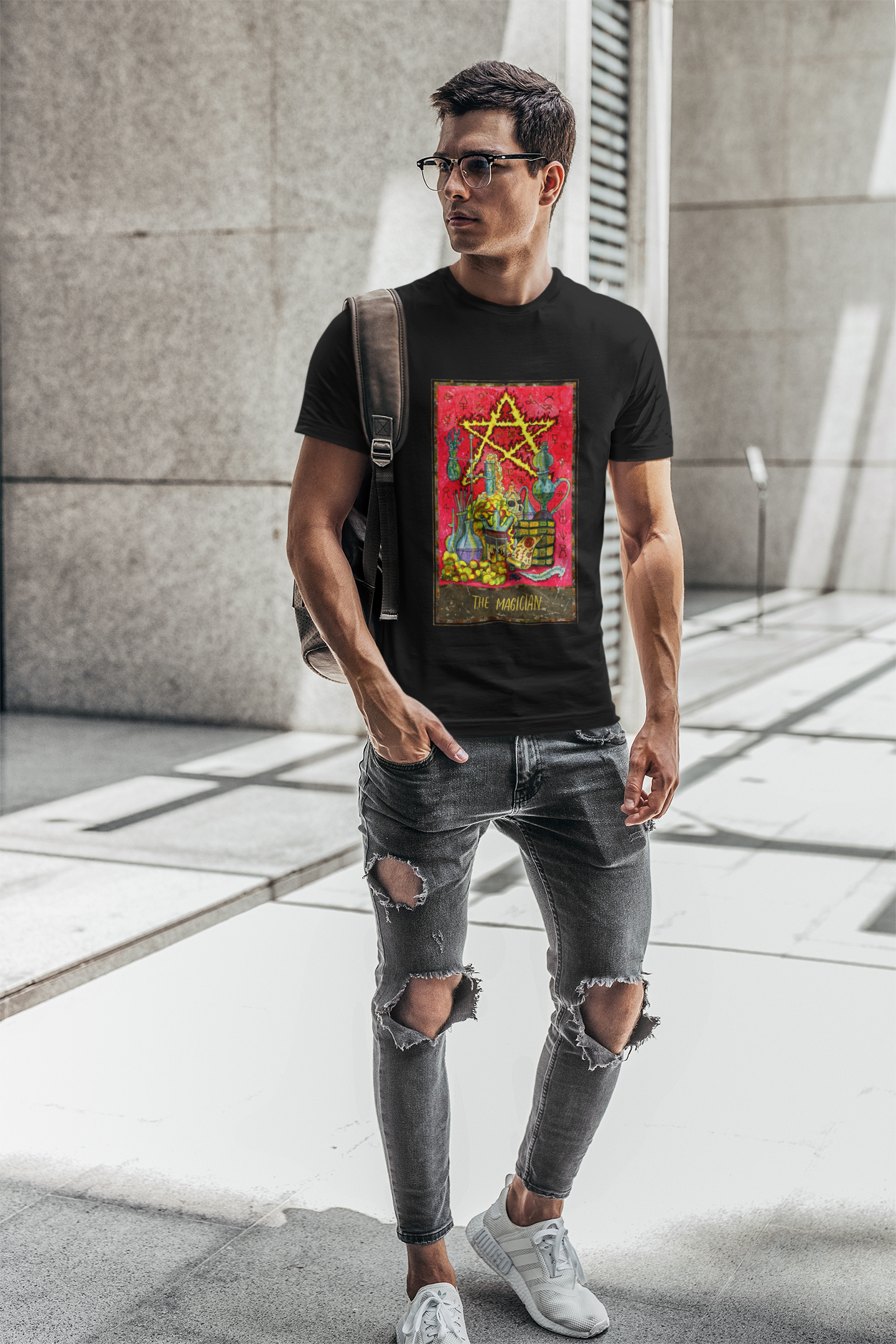 A man with glasses carrying a backpack on his shoulder wearing a black The Magician Tarot Card T-shirt, ripped black jeans, and white shoes