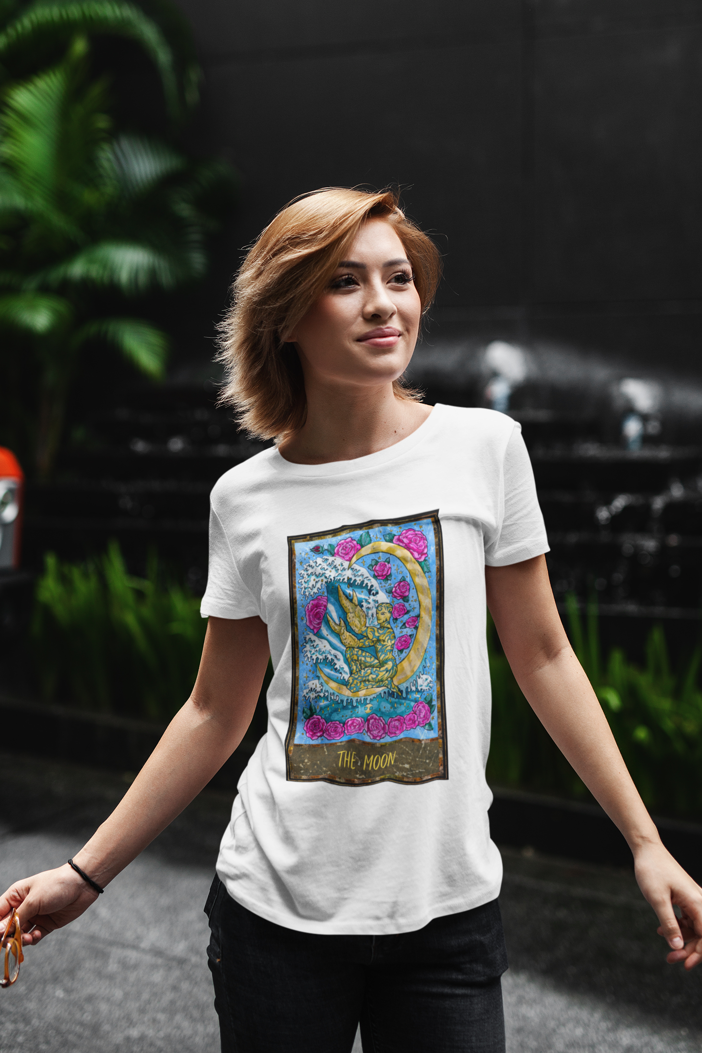 A woman standing against a dark background with some plants wearing a white The Moon Tarot Card T-shirt and black pants